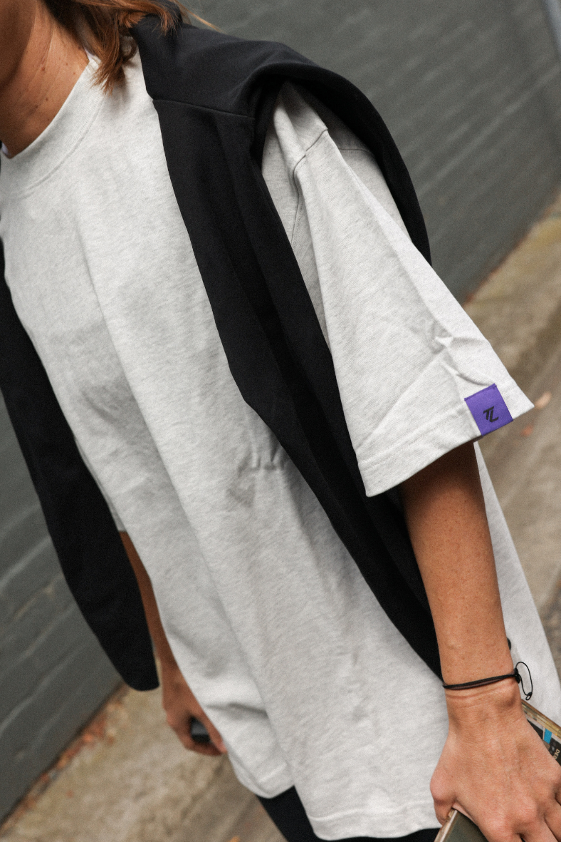 Person wearing a grey t-shirt and a purple tag, standing against a gray wall.