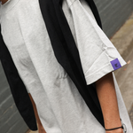 Person wearing a grey t-shirt and a purple tag, standing against a gray wall.