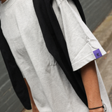 Person wearing a grey t-shirt and a purple tag, standing against a gray wall.