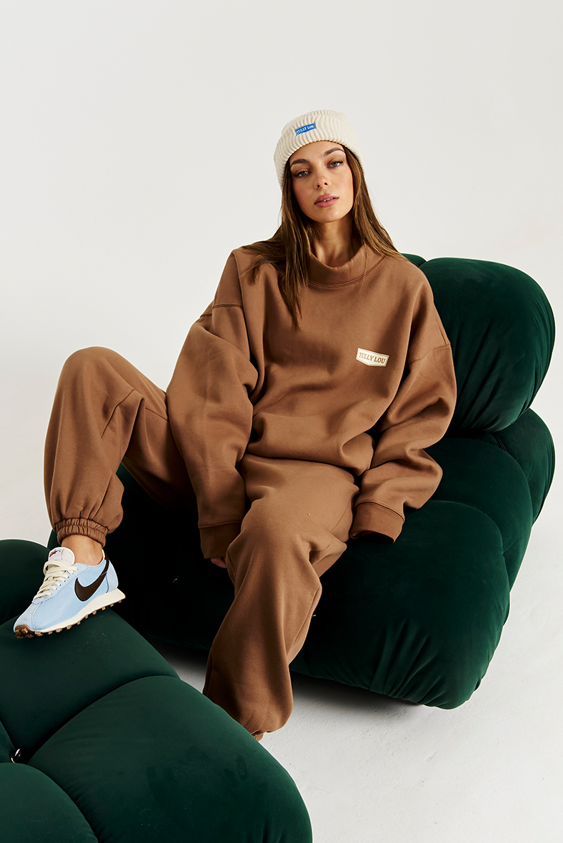 Model on green couch wearing mocha tracksuit heavy fleece set with cream beanie 