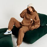 model on green couch wearing brown tracksuit set with cream beanie on and blue and brown sneakers