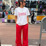 Person wearing a white t-shirt with pink text and red pants standing on a street.